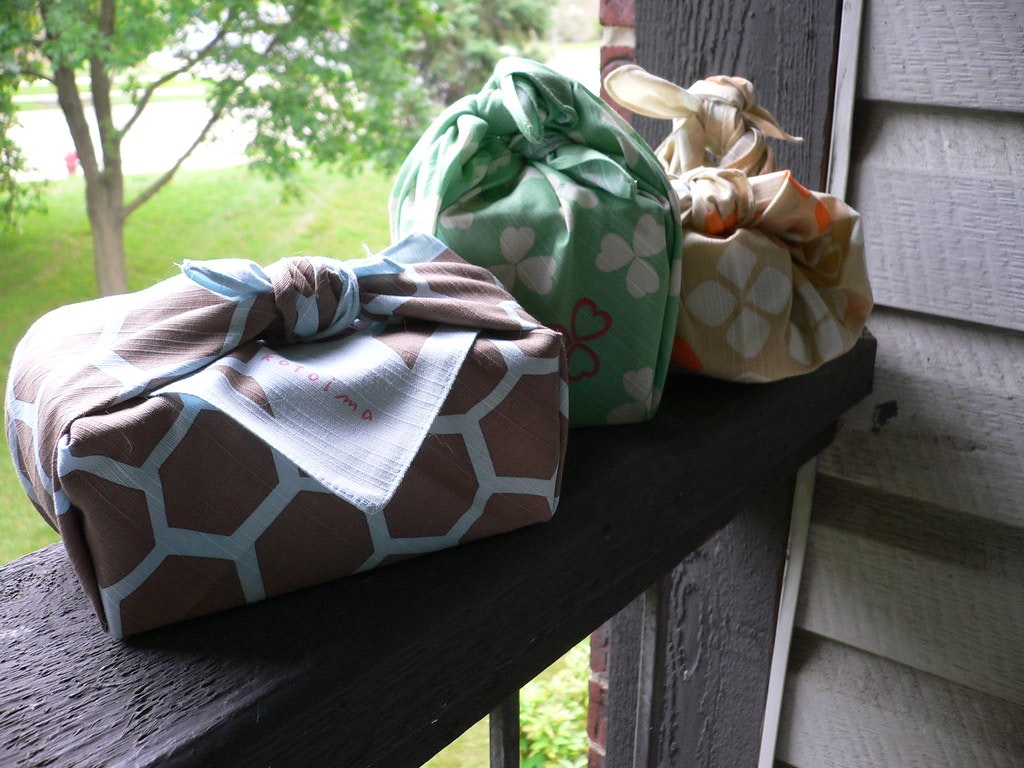 Three fabric-wrapped packages sit on a railing, surrounded by a leafy background.