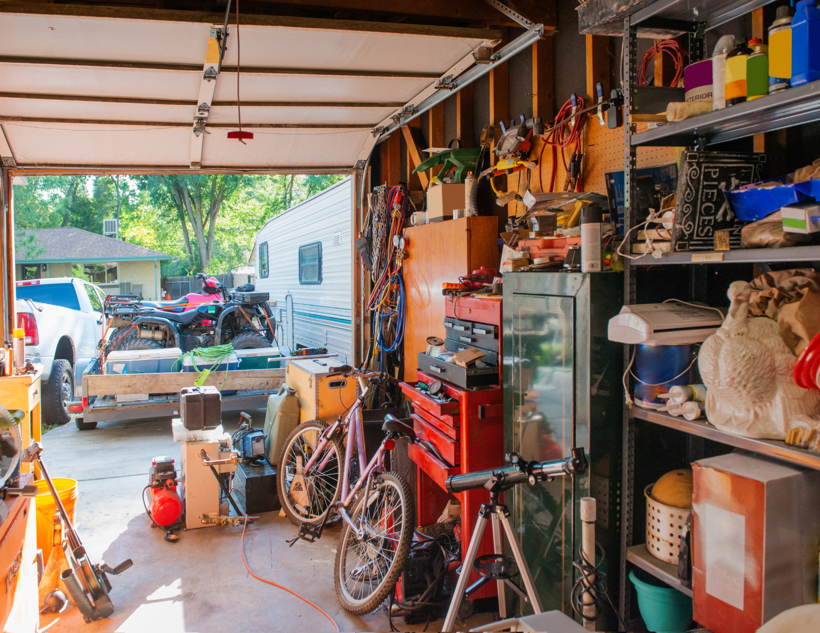 A cluttered garage with tools, bikes, a trailer carrying ATVs, and various equipment.