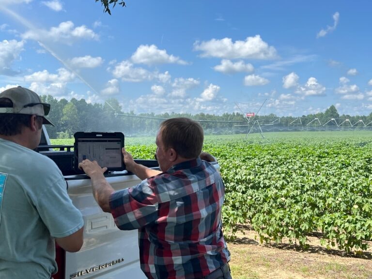 Two people with a laptop by a pickup truck, overlooking an irrigated field on a sunny day.