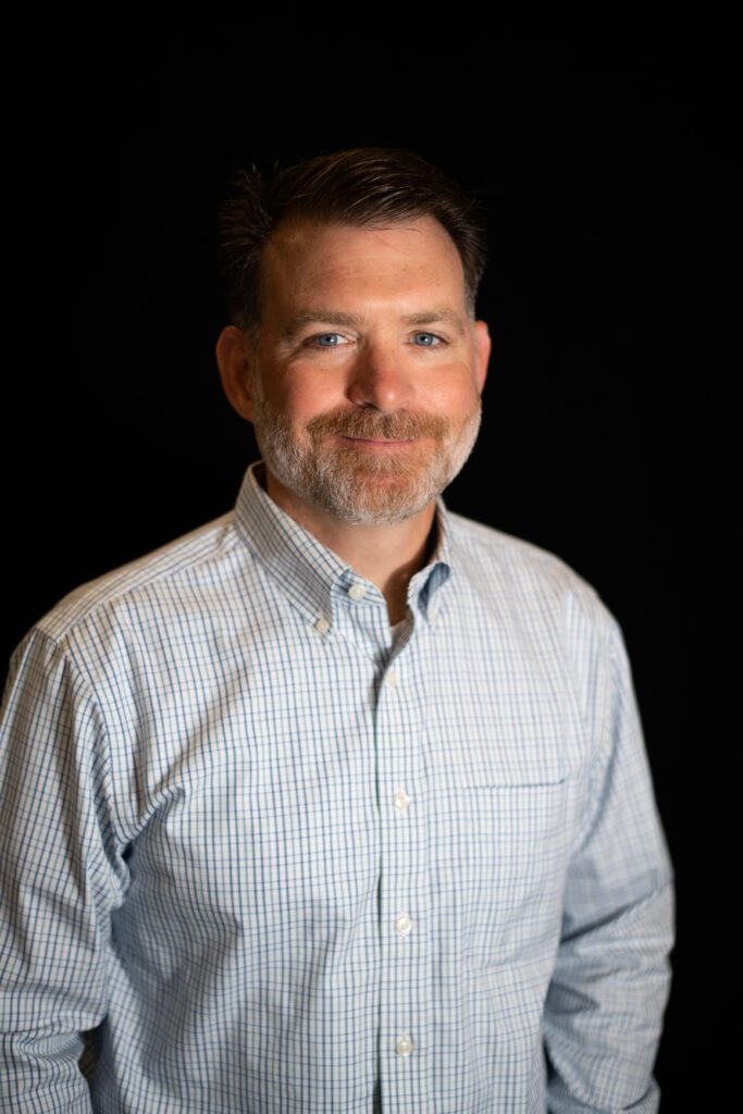 Joyner - A smiling man with a beard wearing a checkered shirt against a black background.