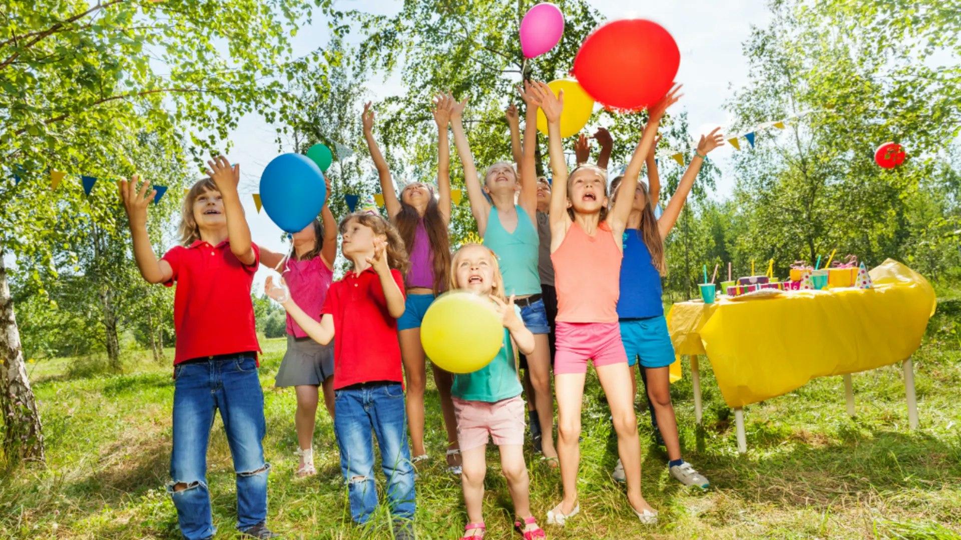 Children playing with balloons at an outdoor party with a table of supplies in the background.