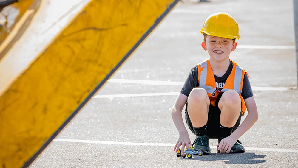 A child in a hard hat and hi-vis vest plays with toy trucks on the ground.