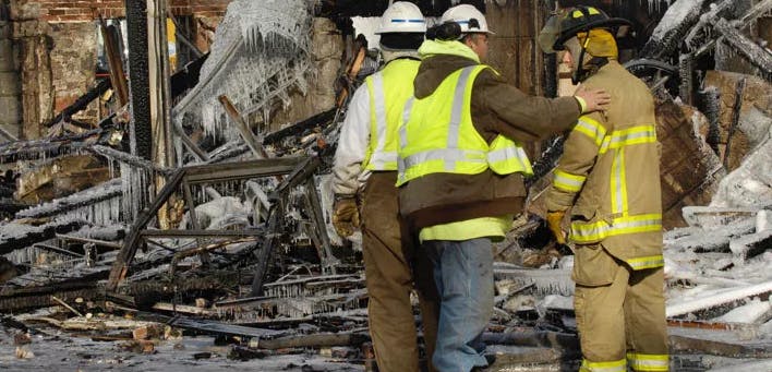 Three people in safety gear and uniforms stand amidst icy debris, possibly after a fire.
