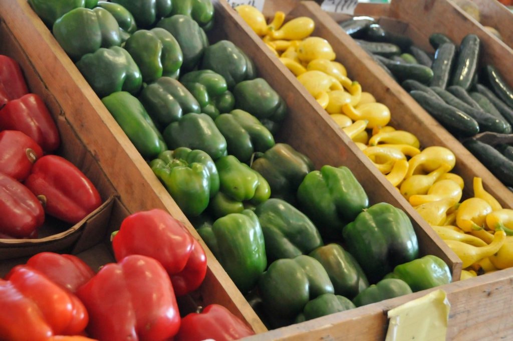 Assorted vegetables displayed in wooden bins: red and green bell peppers, yellow squash, and cucumbers.