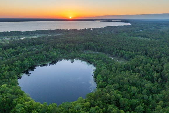 A forested landscape with a circular lake in the foreground and a sunset over a larger body of water in the background.