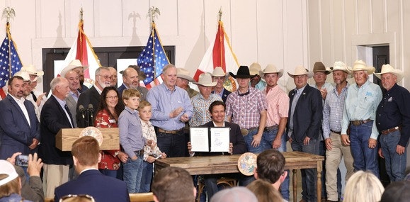 A group of people gather around a person seated at a table holding a document. Flags are in the background.