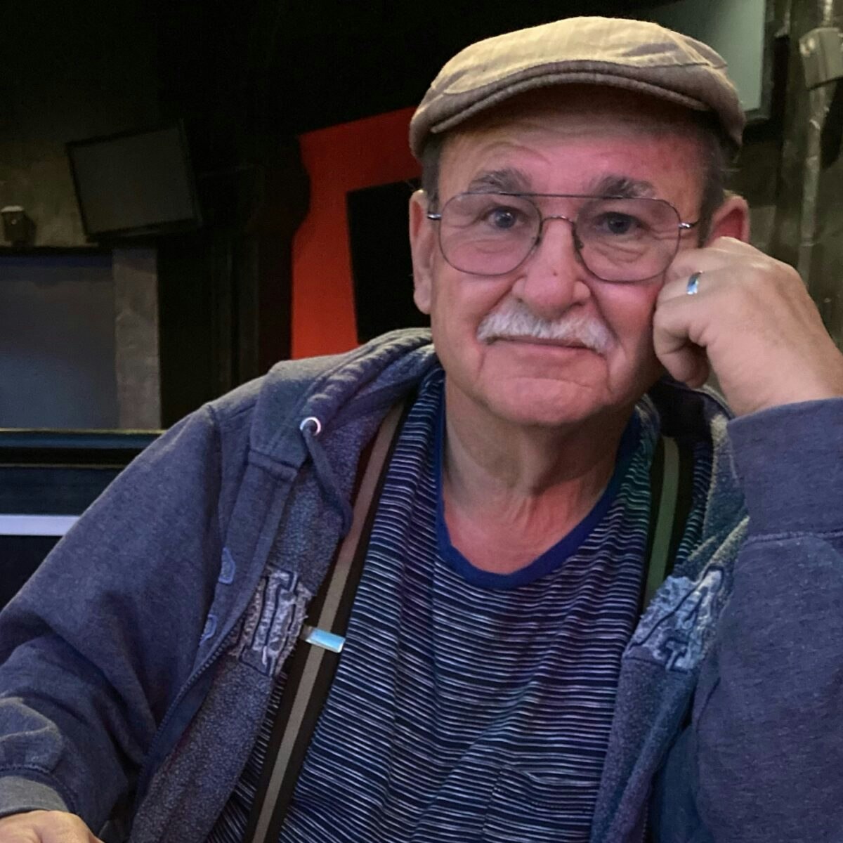 An elderly man wearing a cap and glasses, sitting indoors with a relaxed expression.