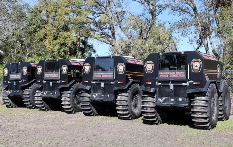 Five off-road vehicles with large tires, marked "FIRE RESCUE," parked on grass.