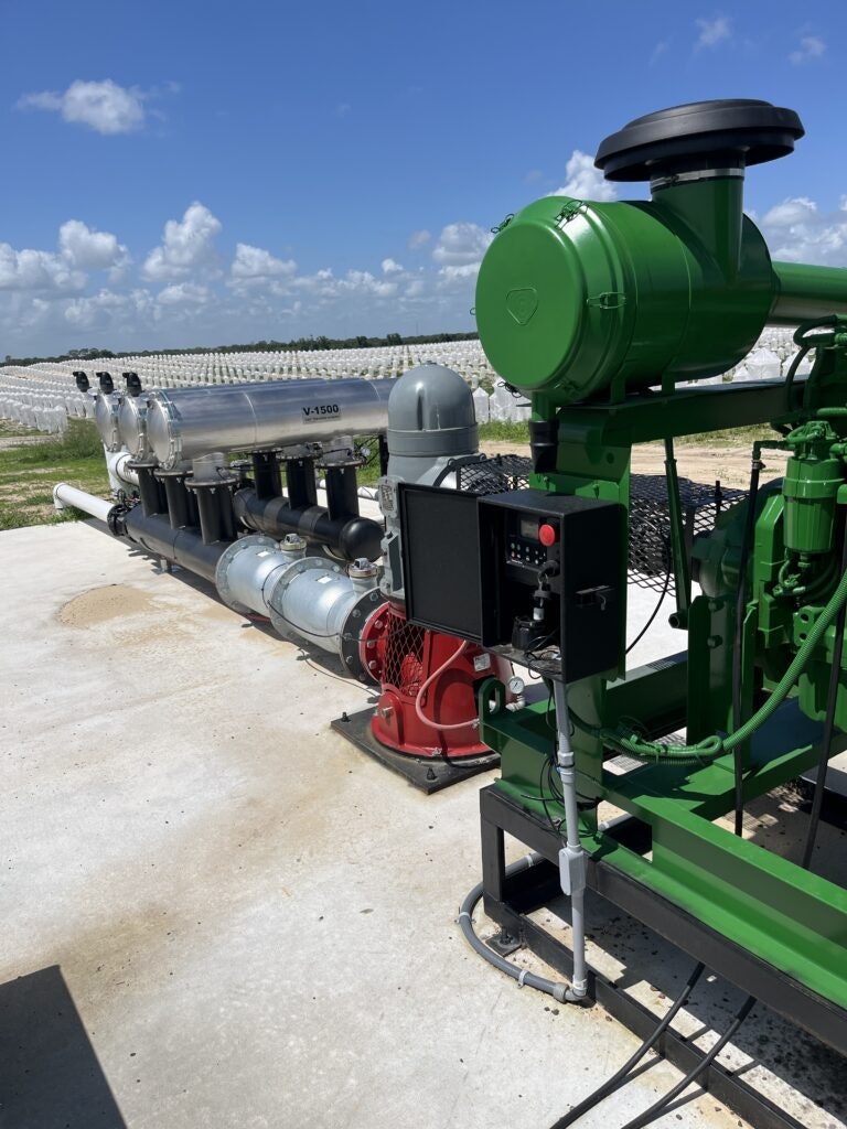 Industrial machinery with pipes and valves, possibly for water treatment, with greenhouse structures in the background under a blue sky.