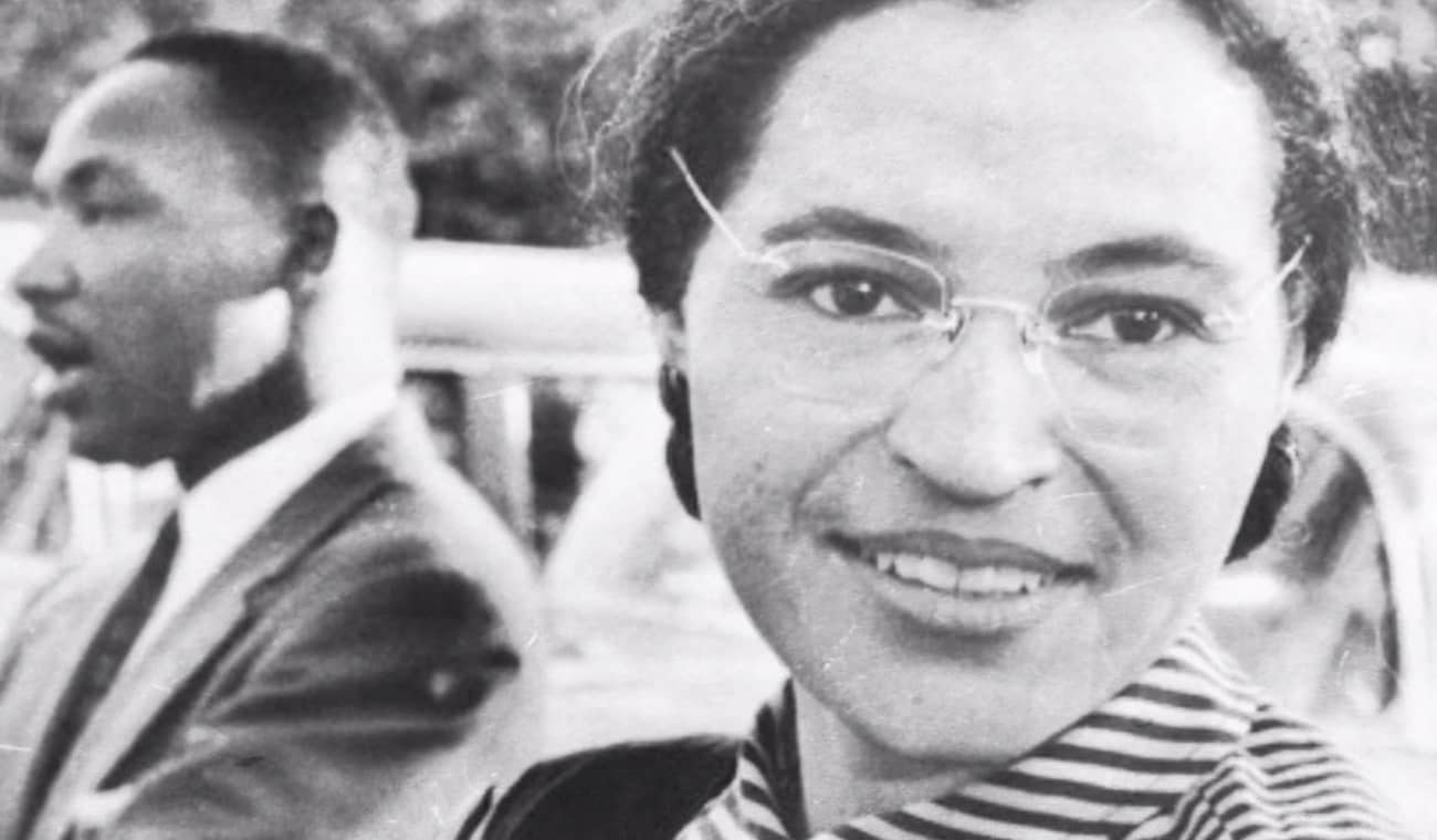 A historic black-and-white photo of a smiling woman in the foreground and a man speaking in the background.