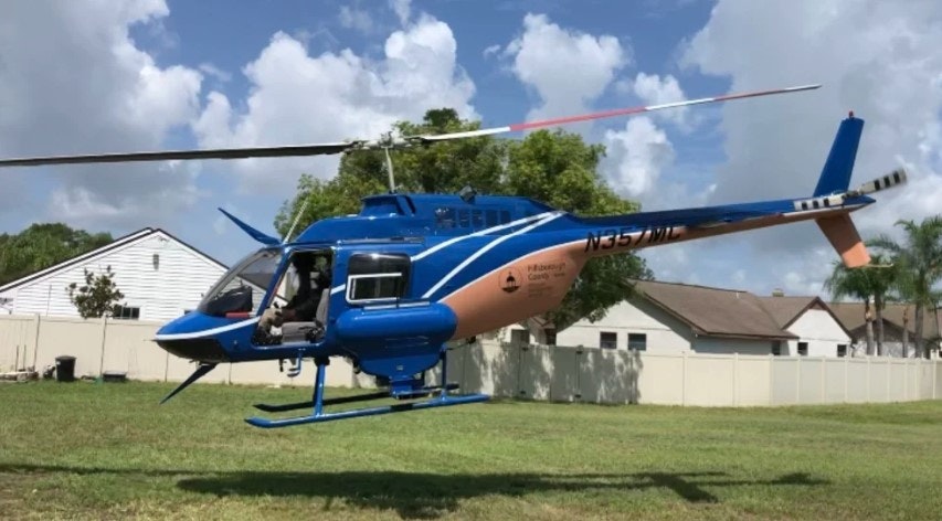 A blue and orange helicopter hovering above a grassy area near residential houses with a pilot inside.