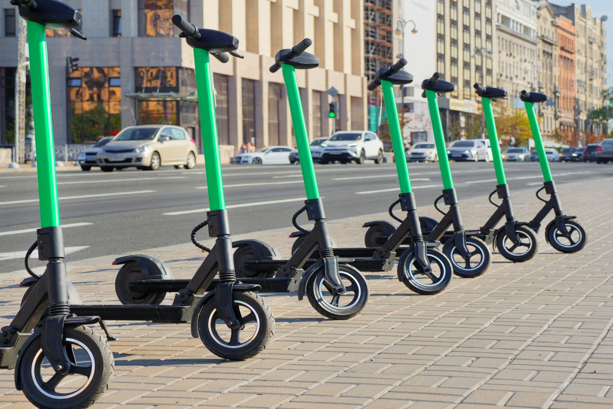 A row of electric scooters on a sidewalk, city street in the background.
