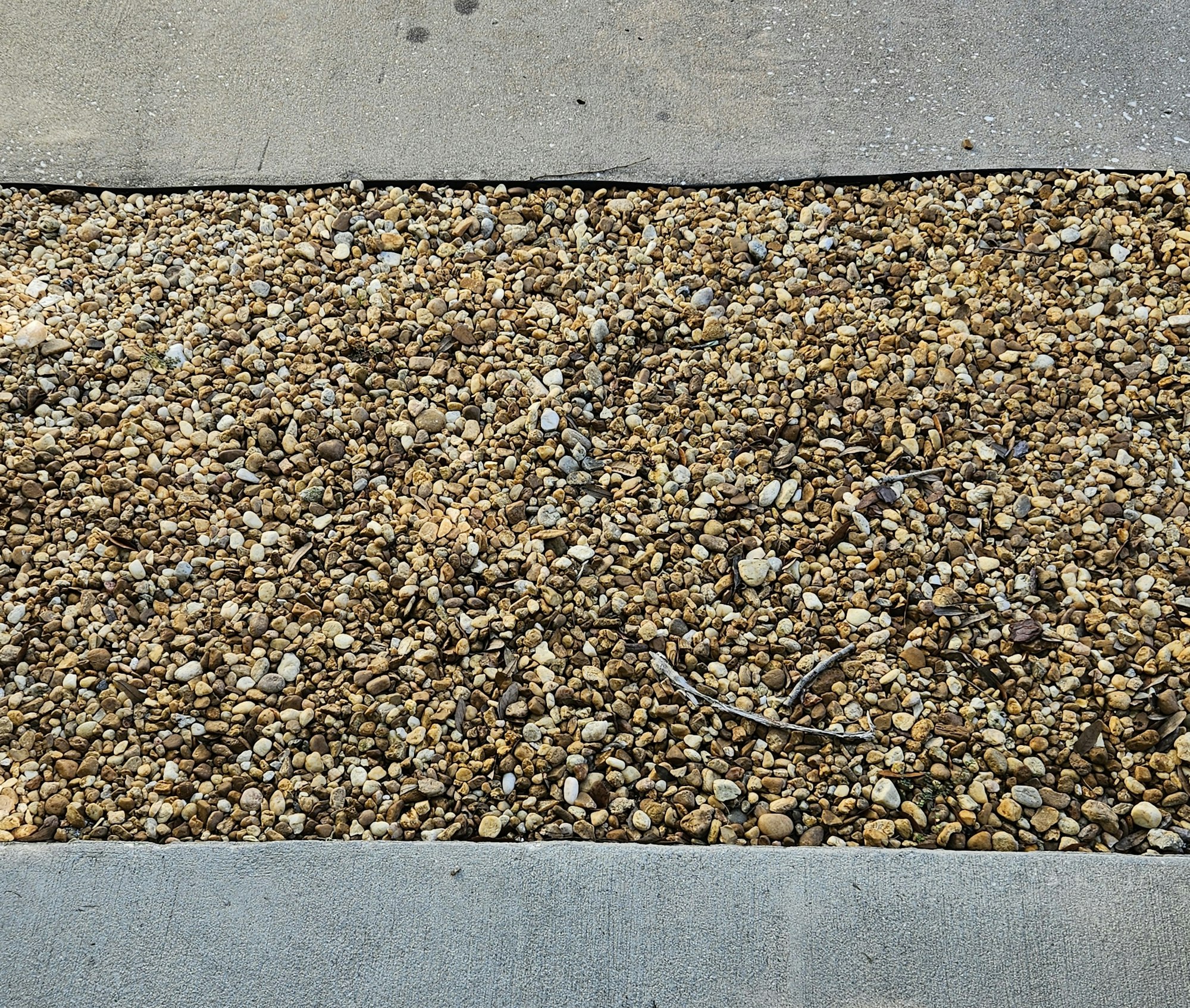 A patch of small, mixed gravel stones next to a concrete surface, creating a permeable barrier for our downspouts..
