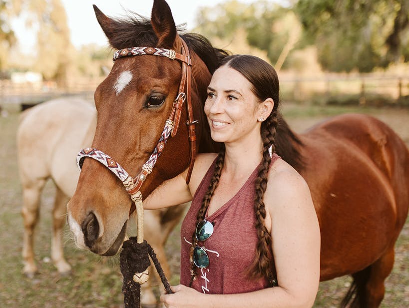 A woman with braided hair stands beside a brown horse, smiling, in an outdoor setting.