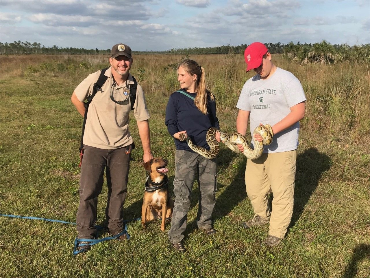 Three people and a dog in a field, one person holding a large snake.