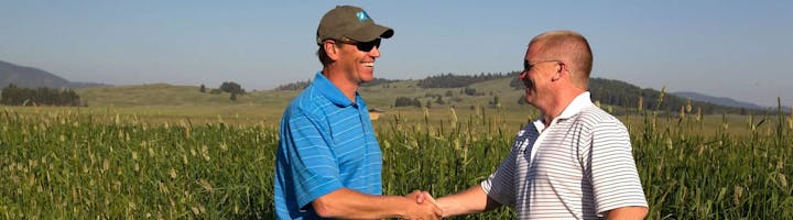 Two men shaking hands in a field with hills in the background.
