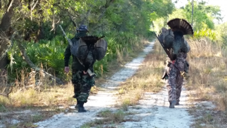 Two people in camo with large turkey fans walking down a trail.