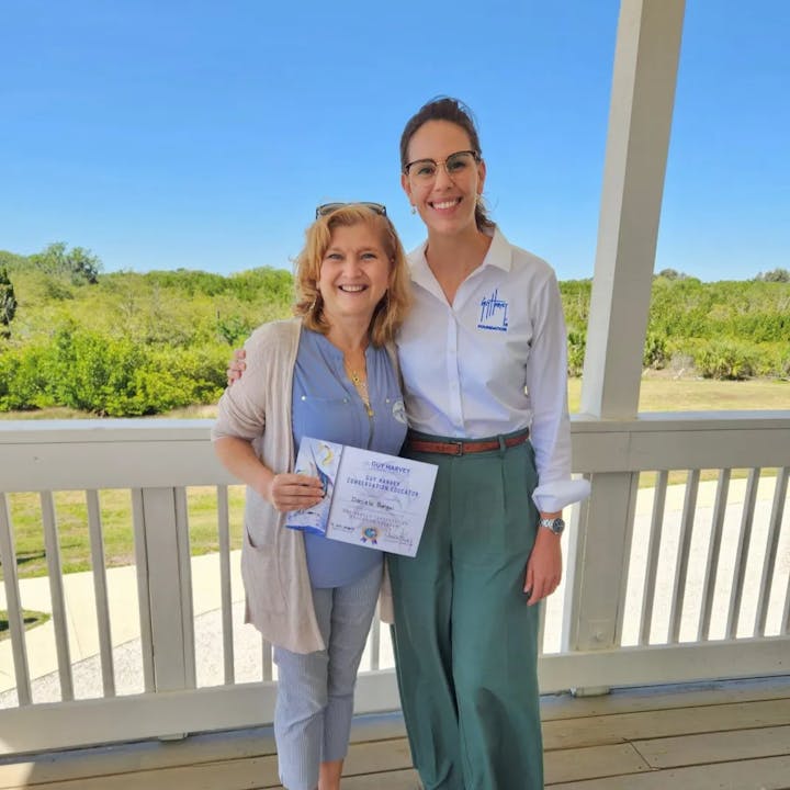 Two smiling women on a porch; one is holding a certificate. Bright day with greenery in the background.