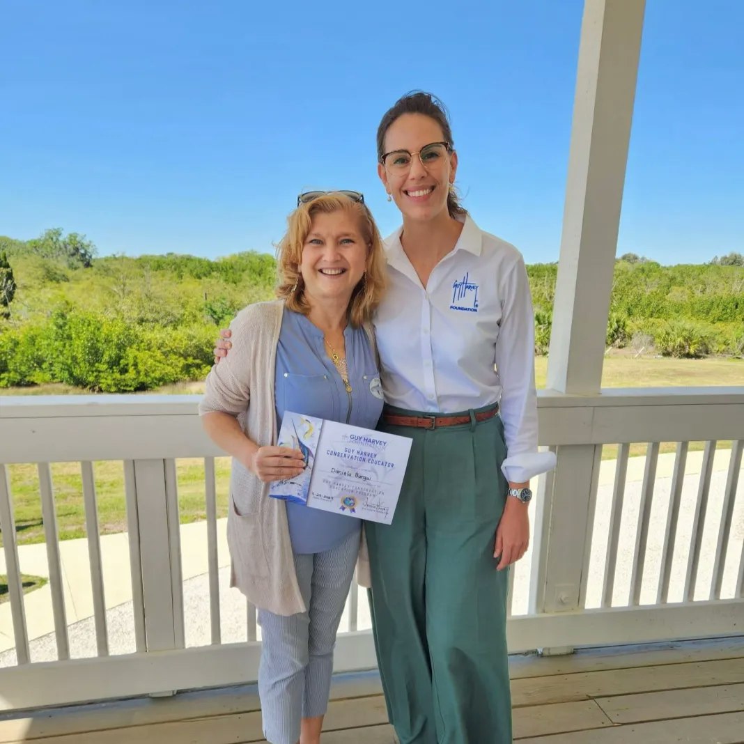 Two smiling women on a porch; one is holding a certificate. Bright day with greenery in the background.