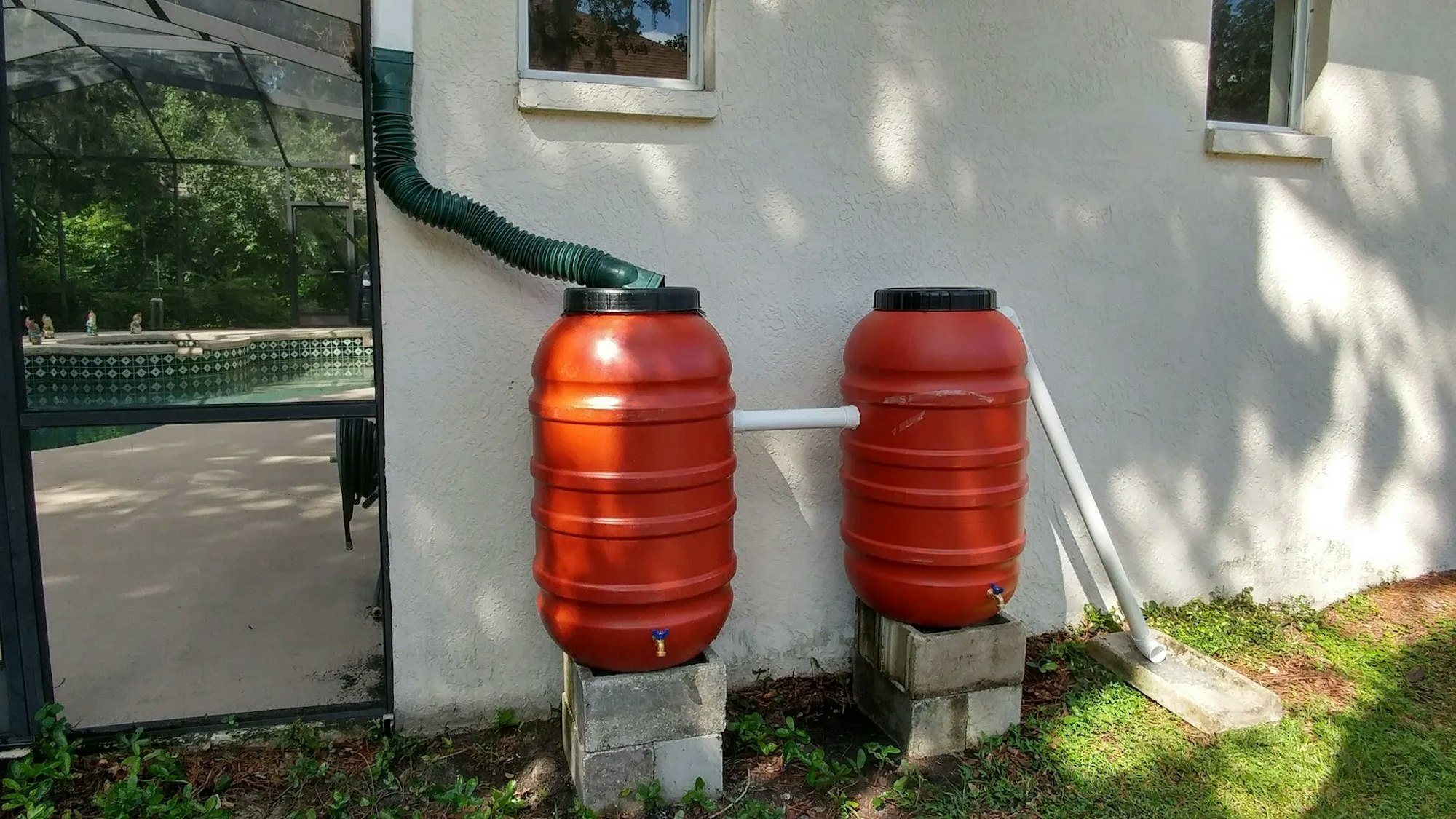 Two red rain barrels are connected to a gutter system, positioned on concrete blocks against a house wall near a pool.