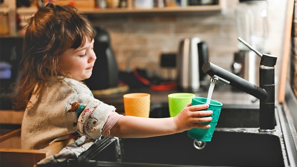 A child filling a cup with water from a kitchen faucet.