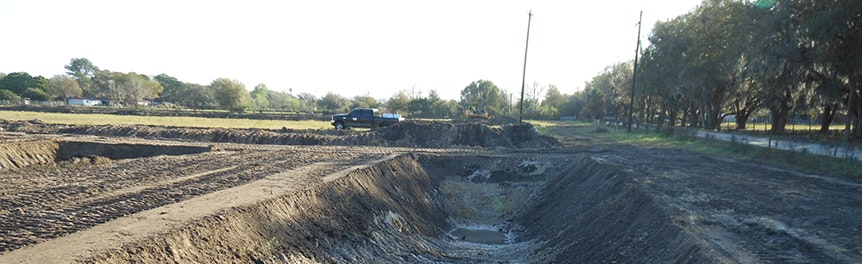 A large, dug-out trench with a truck nearby, set in a rural landscape with trees and fields.