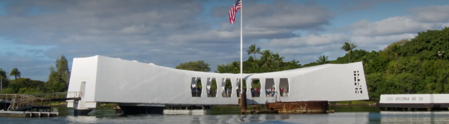 White memorial structure over water with American flag, visitors inside, surrounded by greenery.