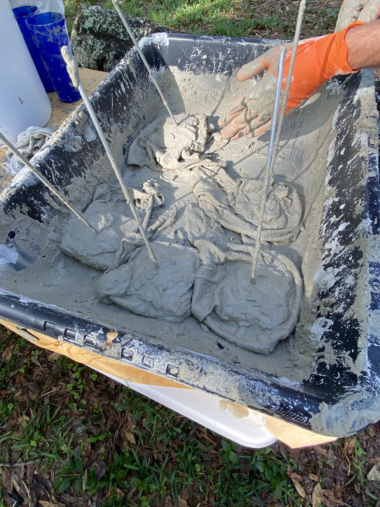 A person with an orange glove is mixing or handling cement or plaster in a tray with metal rods inserted into it.