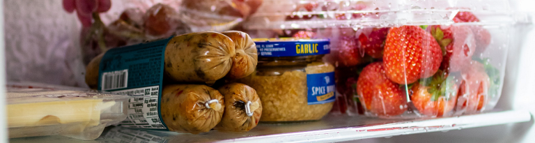 A fridge shelf with cheese, garlic paste, strawberries, and sausages.
