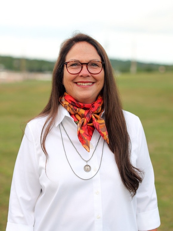 A person with long hair and glasses, wearing a white shirt and a colorful scarf, smiling outdoors.