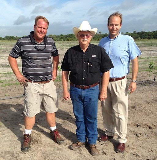 Three men standing outdoors, one in a cowboy hat, on a clear day with sparse vegetation in the background.