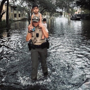 A person in uniform carries a child on their shoulders through a flooded street.
