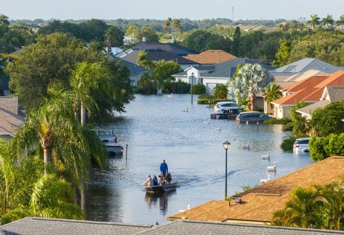 A flooded residential street with a small boat carrying people, surrounded by partially submerged cars and houses.