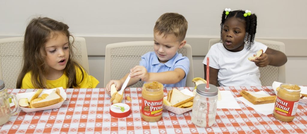 Three children are at a table with peanut butter jars, bread, and apple slices, seemingly making sandwiches.