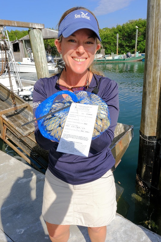 A person holding a blue net bag of clams with a tag, standing by the water.