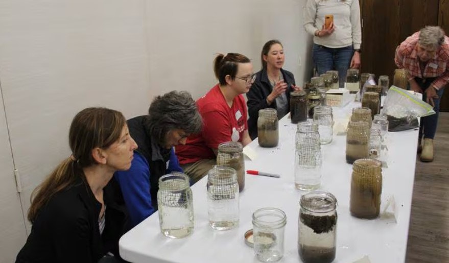 A group is gathered around a table with jars of water and soil samples.