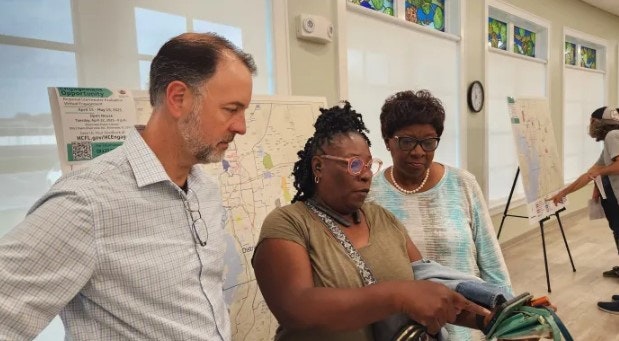 Three people looking at something together, with maps on easels in the background.