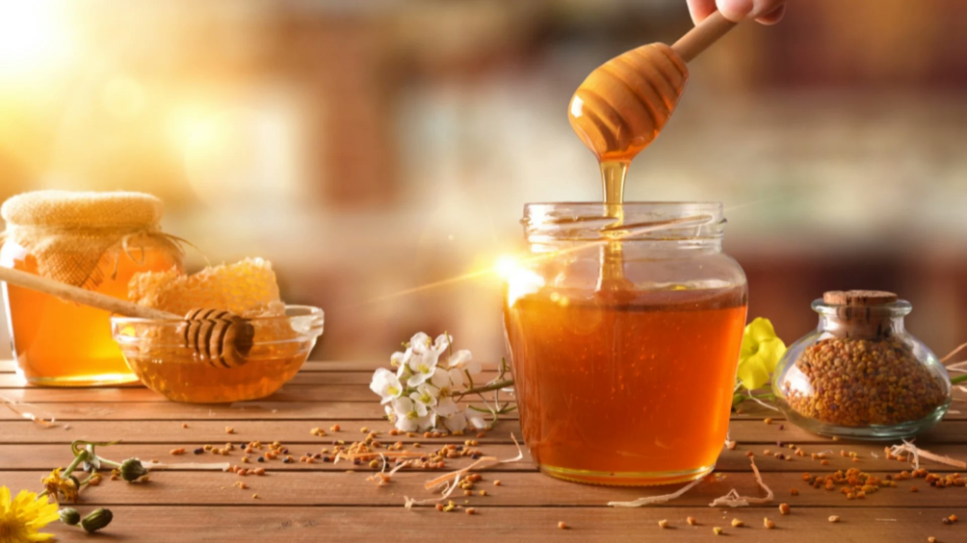A jar of honey with a dipper, honeycomb, flowers, pollen grains, and a warm sunlight backdrop.