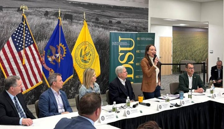 A panel of people sits at a table with flags behind, one woman is speaking into a microphone. North Dakota State University banner visible.