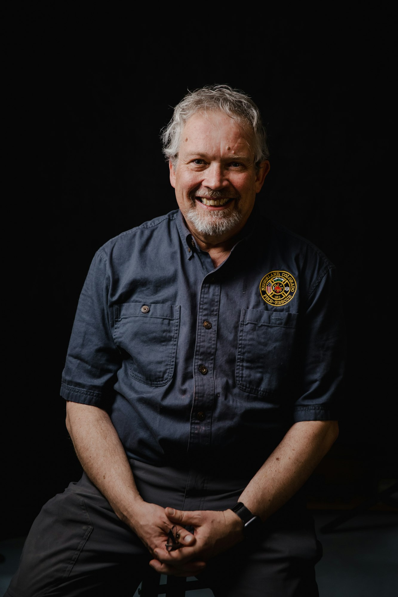 A smiling man in a blue shirt sits against a black background, hands clasped in front of him, exuding a friendly demeanor.