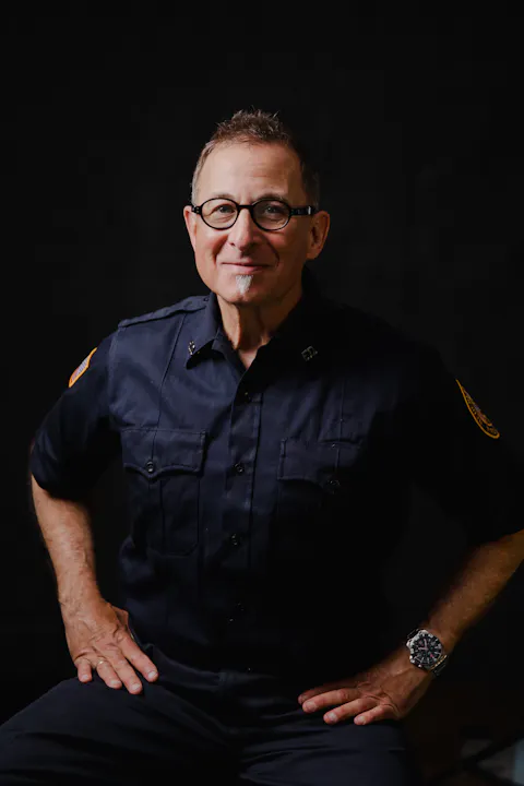 Man in glasses wearing a dark uniform with patches, smiling at the camera against a black background.