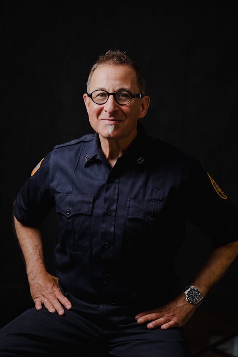 Man in glasses wearing a dark uniform with patches, smiling at the camera against a black background.