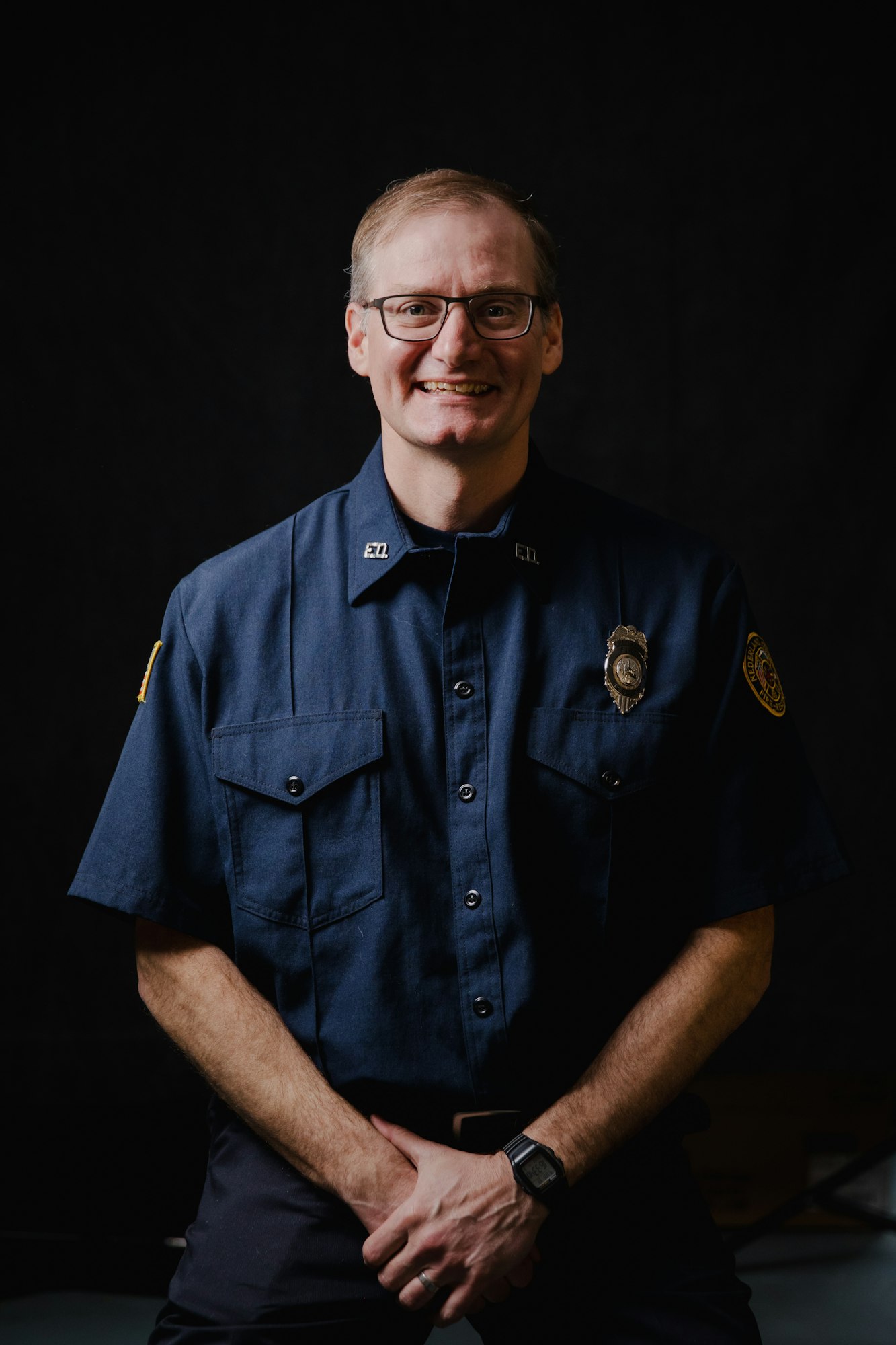 A smiling person in a dark uniform, possibly a firefighter or EMT, with a badge and glasses, against a black background.