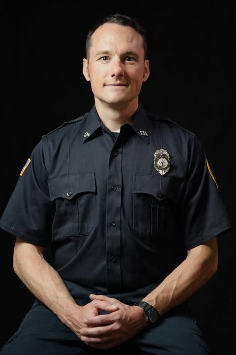 A man in a police uniform sitting with crossed hands against a black background.