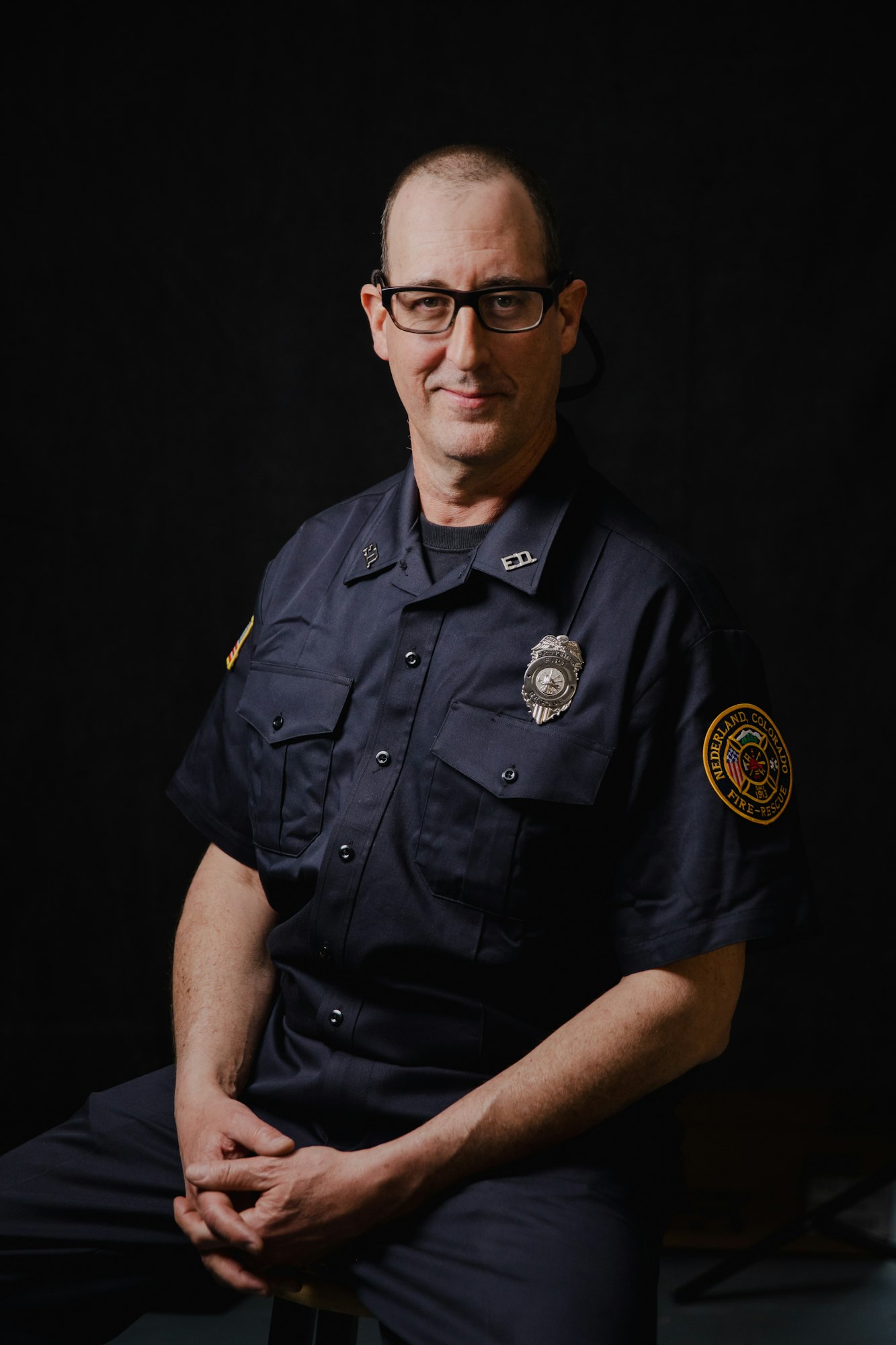 A man in firefighter uniform sitting, with a badge on his shirt and a patch on his sleeve.