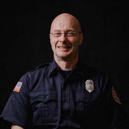 A smiling bald man wearing a police uniform with a badge and an American flag on his sleeve, against a dark background.