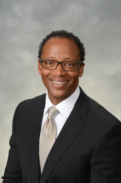 The image features a smiling man in a suit with glasses, set against a light background.