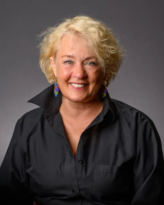 The image features a smiling woman with short, curly blonde hair, wearing a black shirt and colorful earrings against a gray background.