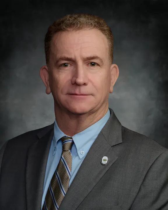 The image shows a professional portrait of a man in a suit, with a blue shirt and striped tie. A badge is visible on his lapel.