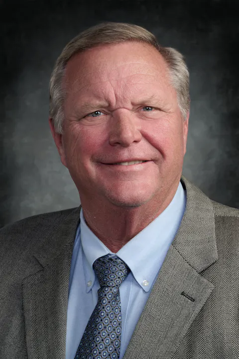 The image features a smiling man in a suit and tie against a dark background. He has short hair and is looking directly at the camera.